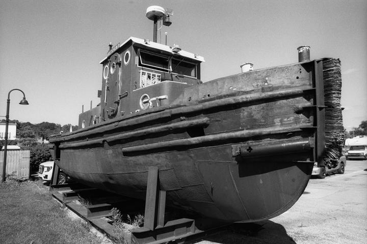 A large tugboat is dry-docked on land next to a street light and parked vehicles.