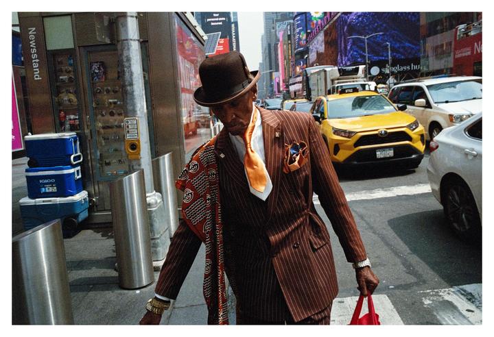 A man in a pinstripe suit and bowler hat walks past a newsstand and yellow taxi in New York.