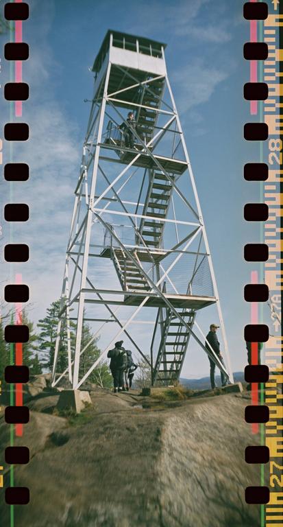 A fire tower stands tall on a rocky hill with several people around its base and one person on its stairs.
