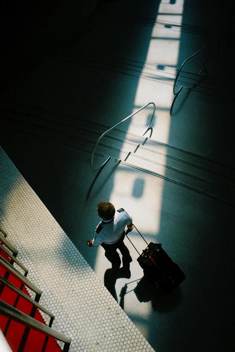 A man in a pilot uniform pulls a rolling suitcase across a tiled floor with strong shadows and light.