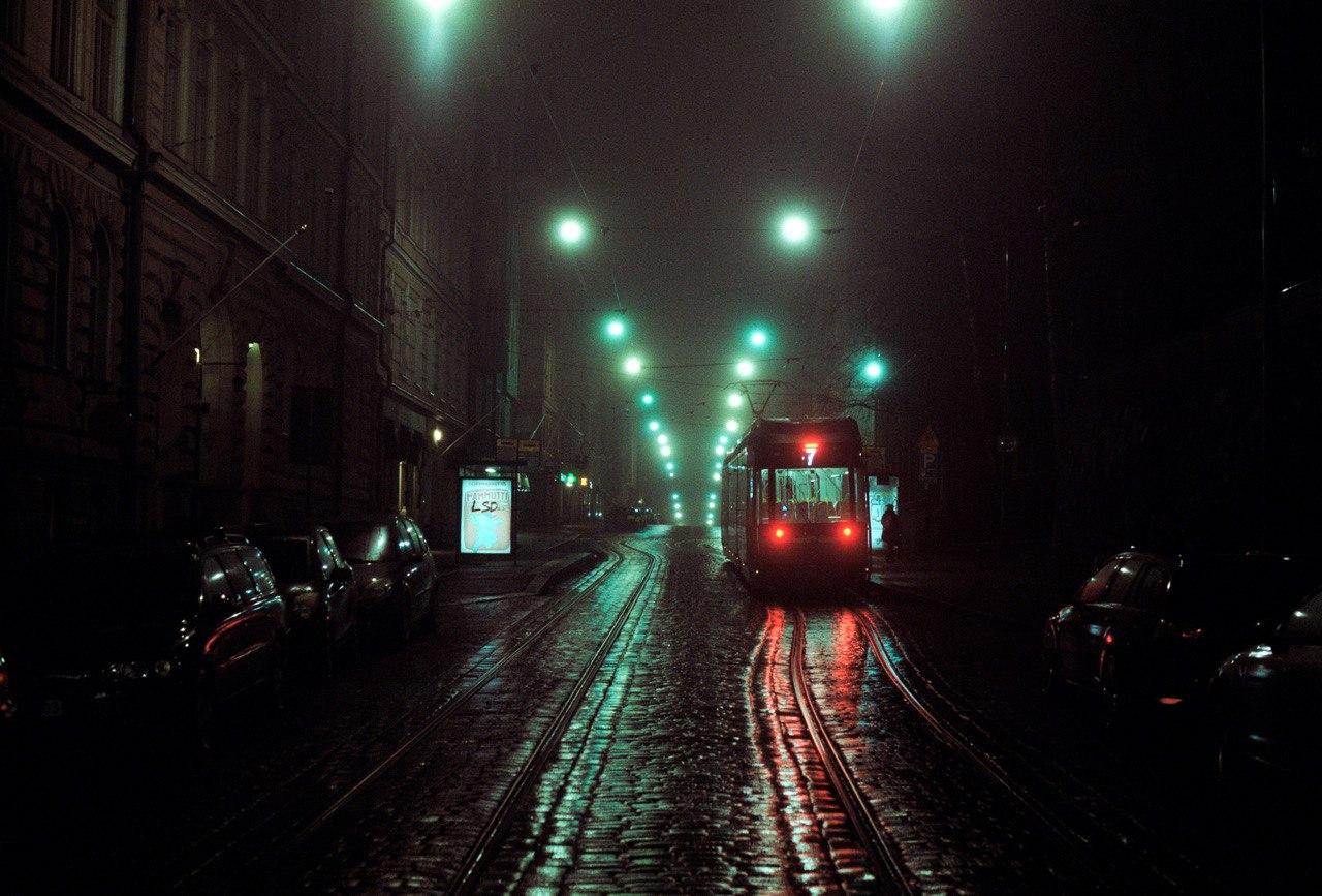 A tram with red lights approaches on a wet street at night, with green streetlights illuminating the fog.