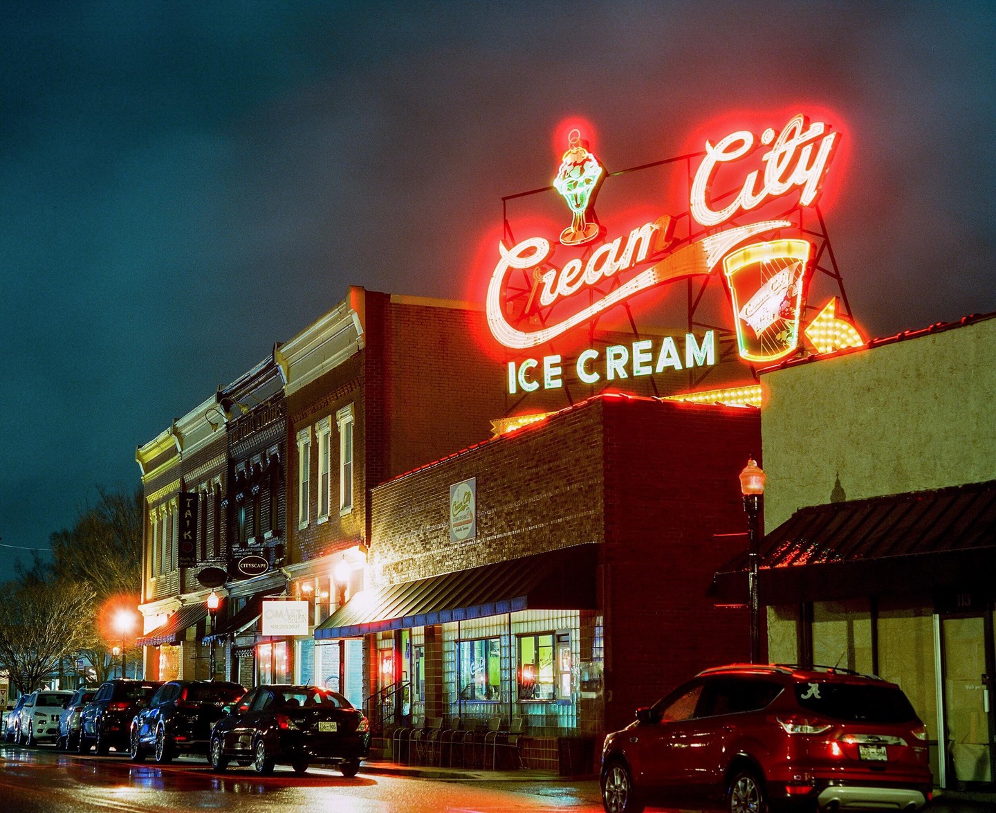 A large neon sign for Cream City Ice Cream illuminates a street with parked cars and brick buildings at night.
