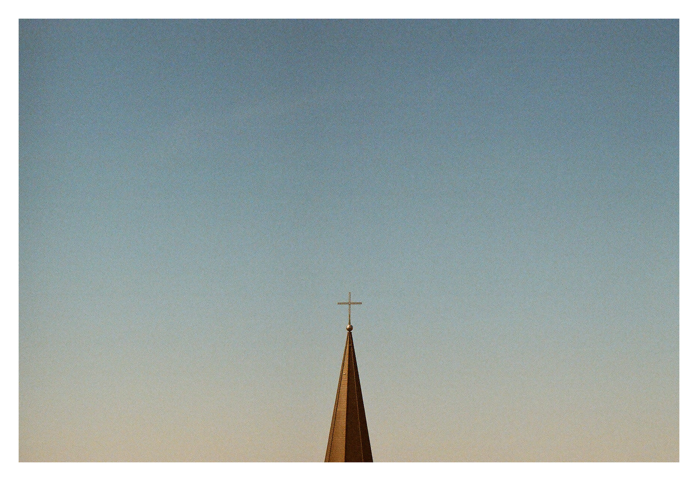 A church spire with a cross on top is silhouetted against a clear sky.