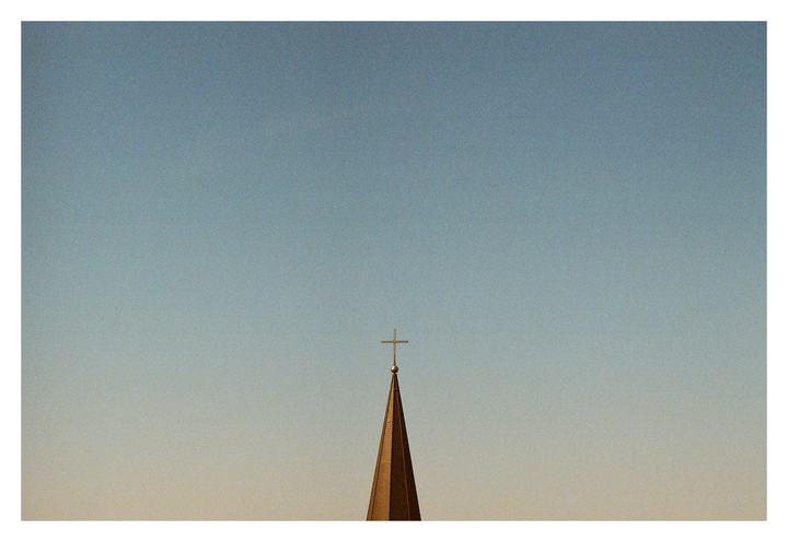 A church spire with a cross on top is silhouetted against a clear sky.