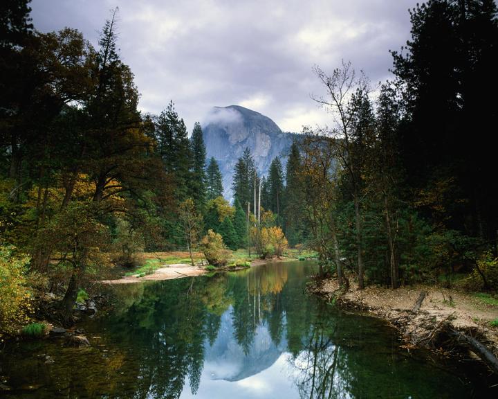 A river reflects a mountain and trees under a cloudy sky.