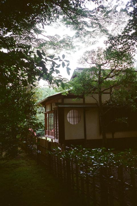 A traditional Japanese house with a circular window is surrounded by lush green foliage and a wooden fence.