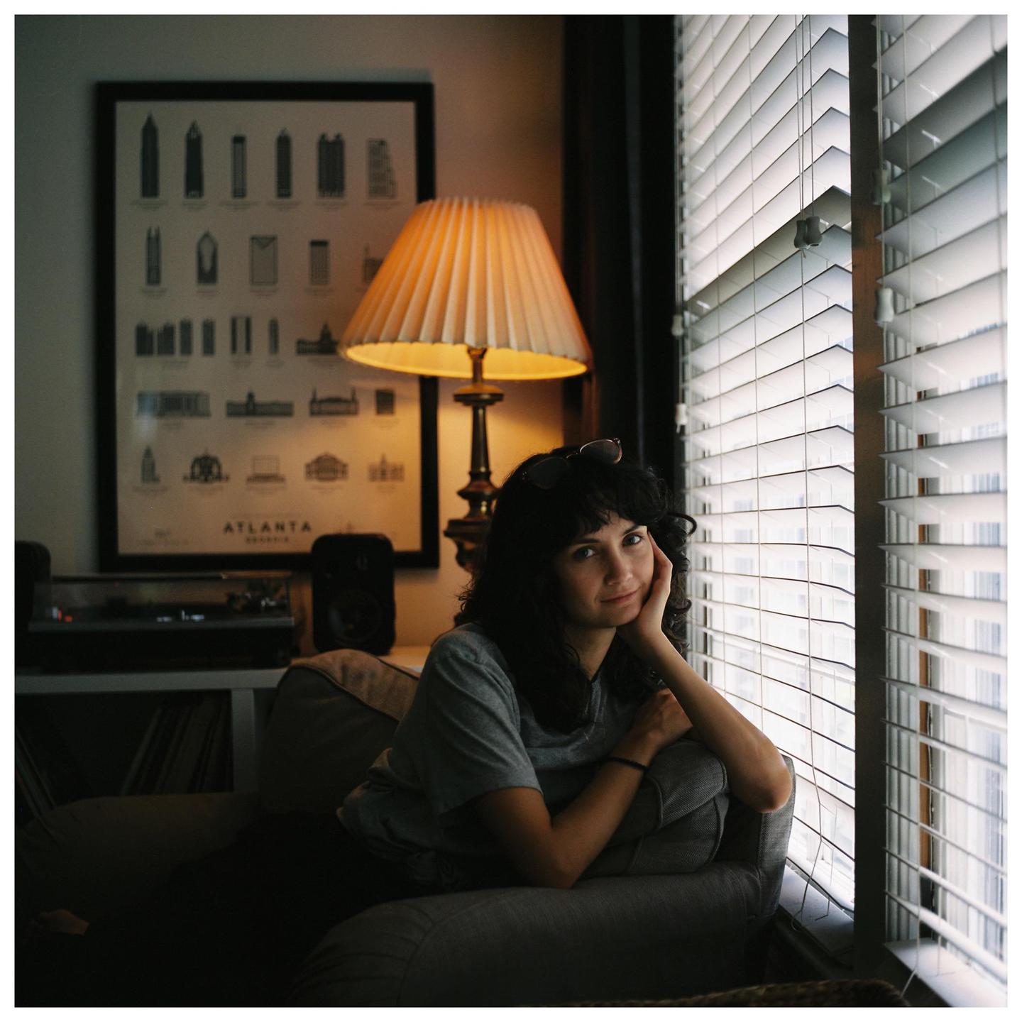 A woman sits on a sofa next to a window with blinds, with a lamp and a framed poster of Atlanta in the background.