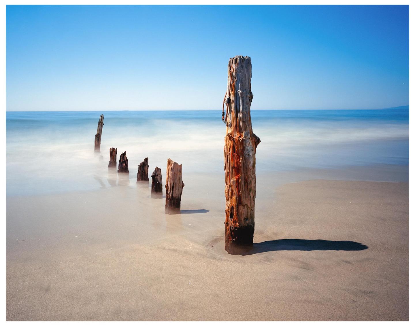 A line of wooden posts extends into the ocean from a sandy beach under a clear blue sky.