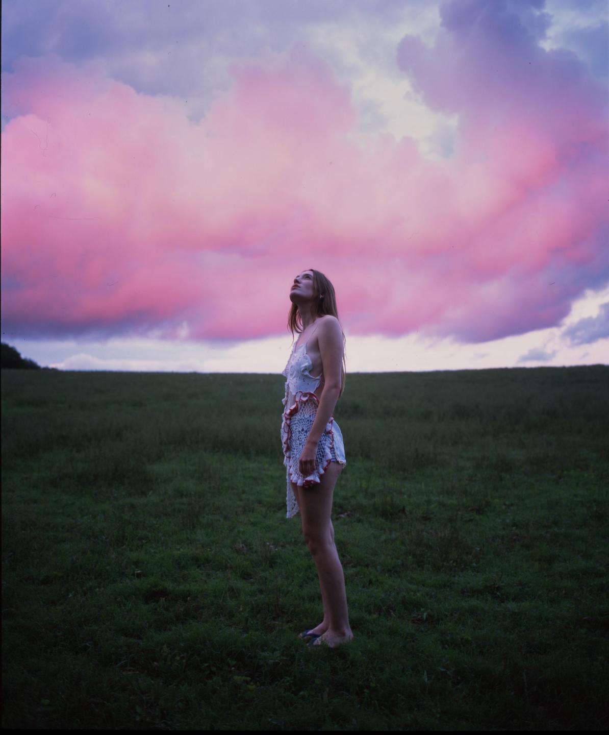 A woman stands in a field looking up at a sky filled with pink clouds.