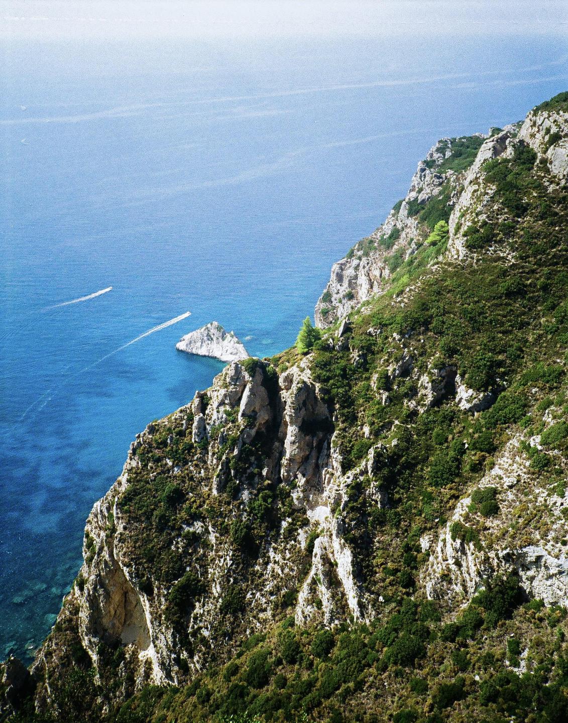 A high-angle view shows a rocky, green cliffside descending into a blue sea with two boats and a small island.