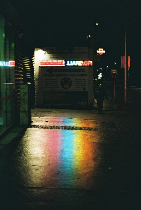 A rainbow reflection from a neon sign illuminates a wet street at night as a person walks by.