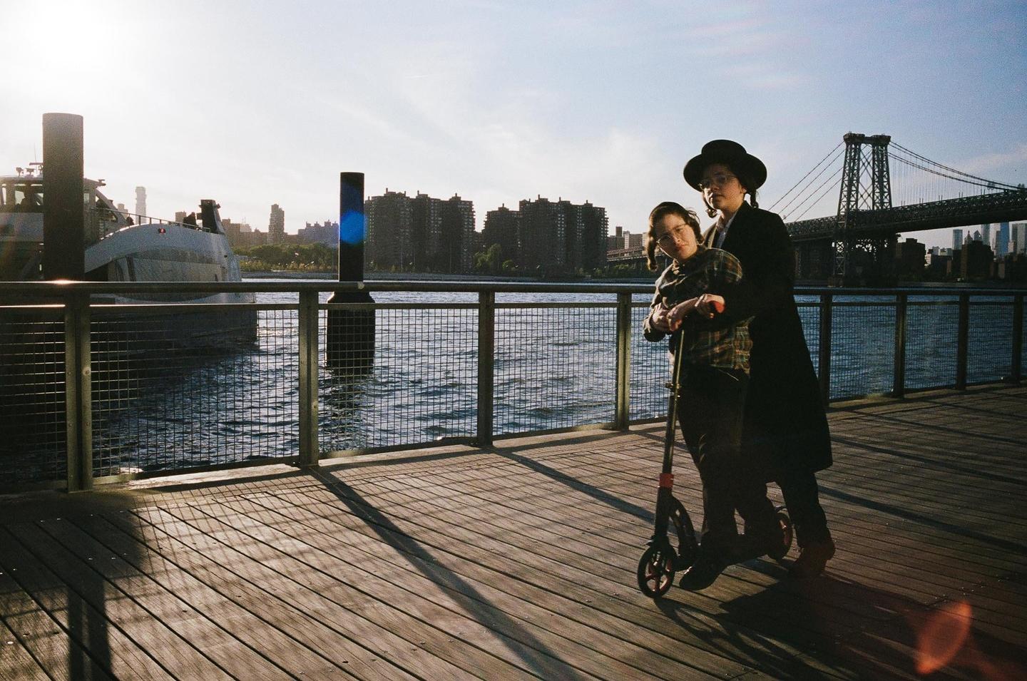 A couple stands on a wooden pier next to a body of water with a bridge and city skyline in the background.