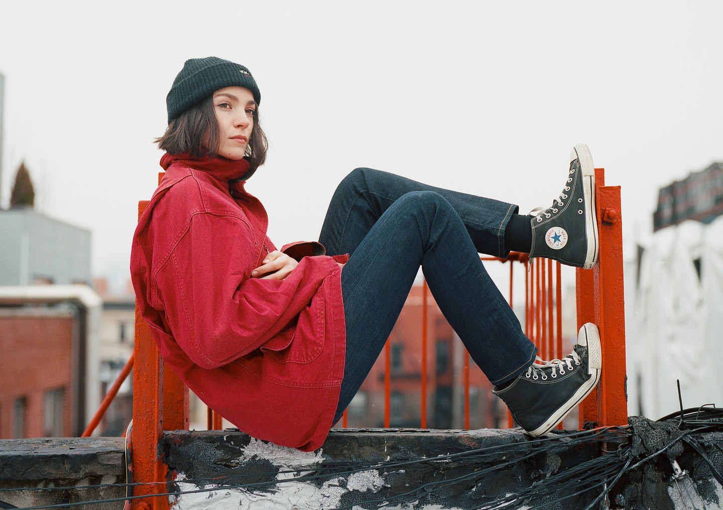 A woman in a red jacket and jeans sits on a rooftop ledge with her legs propped up.