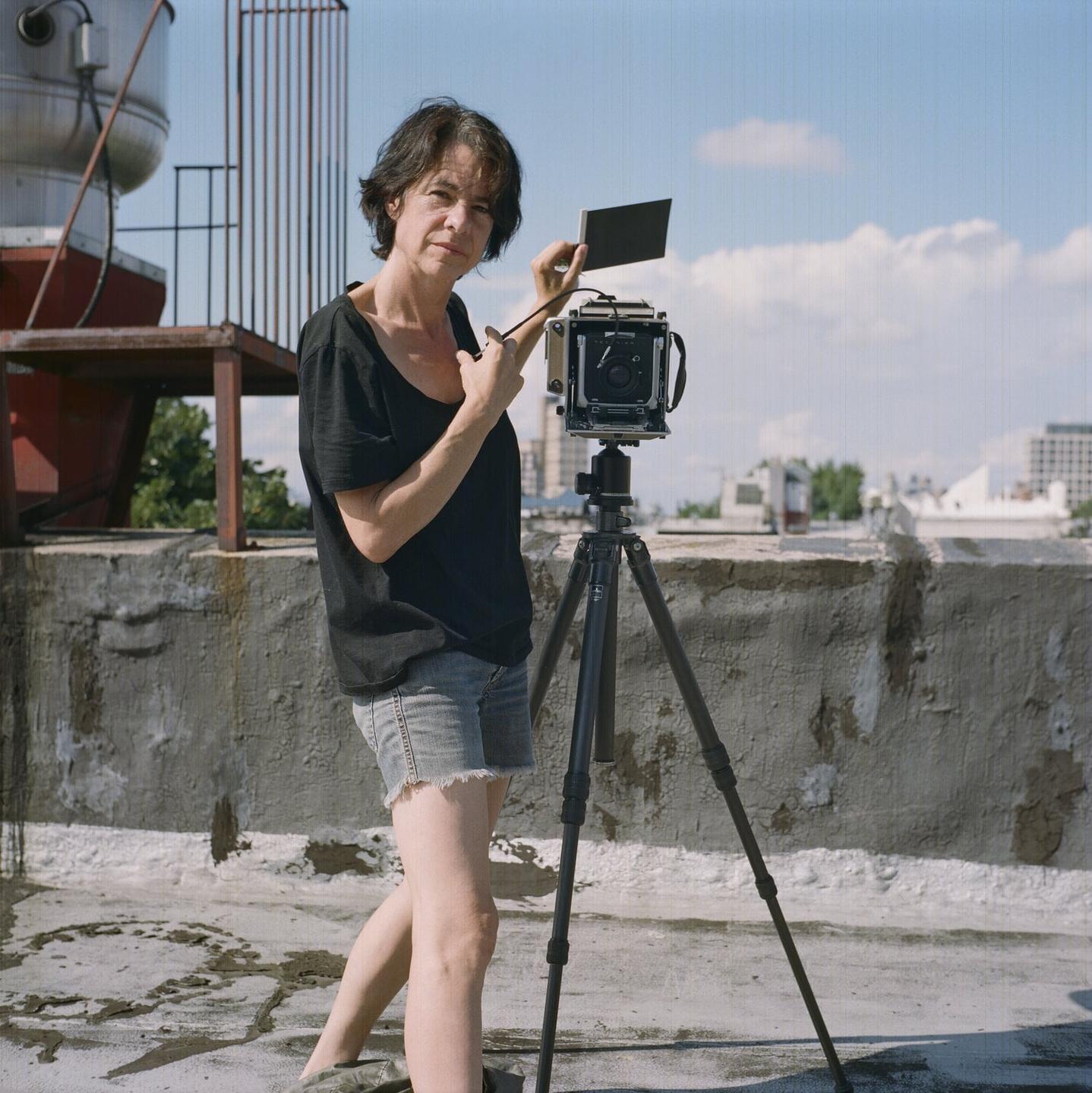 A woman stands on a rooftop next to a large format camera on a tripod, holding a dark slide.