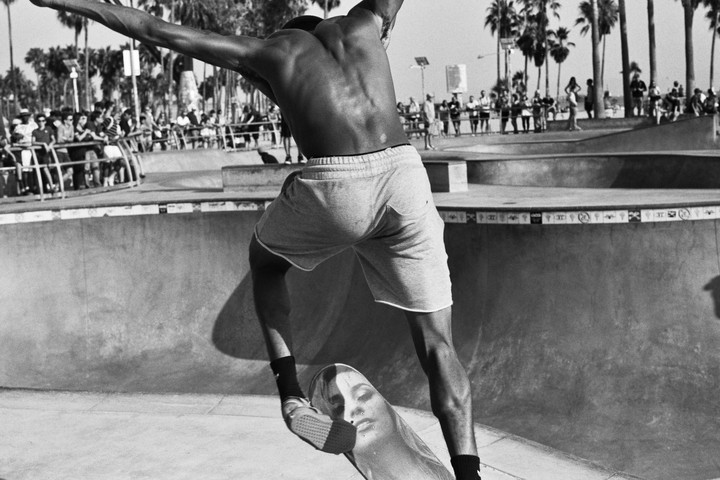 A man in shorts balances on a skateboard in a skate park with palm trees and spectators in the background.