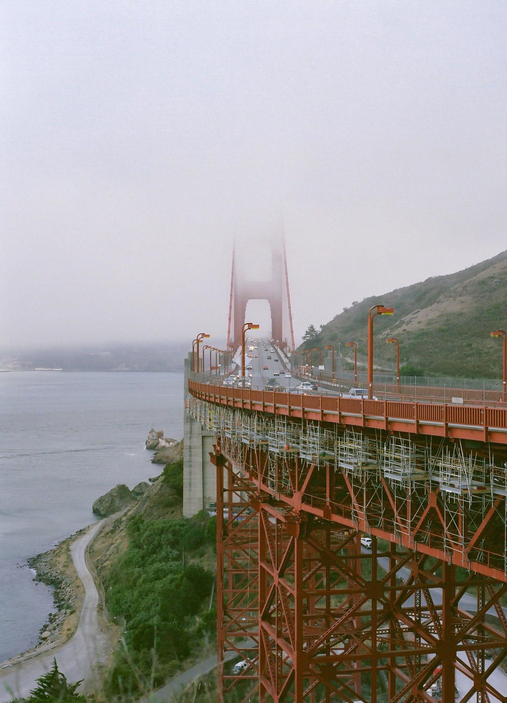 The Golden Gate Bridge is shrouded in fog, with cars driving across its span and scaffolding visible underneath.