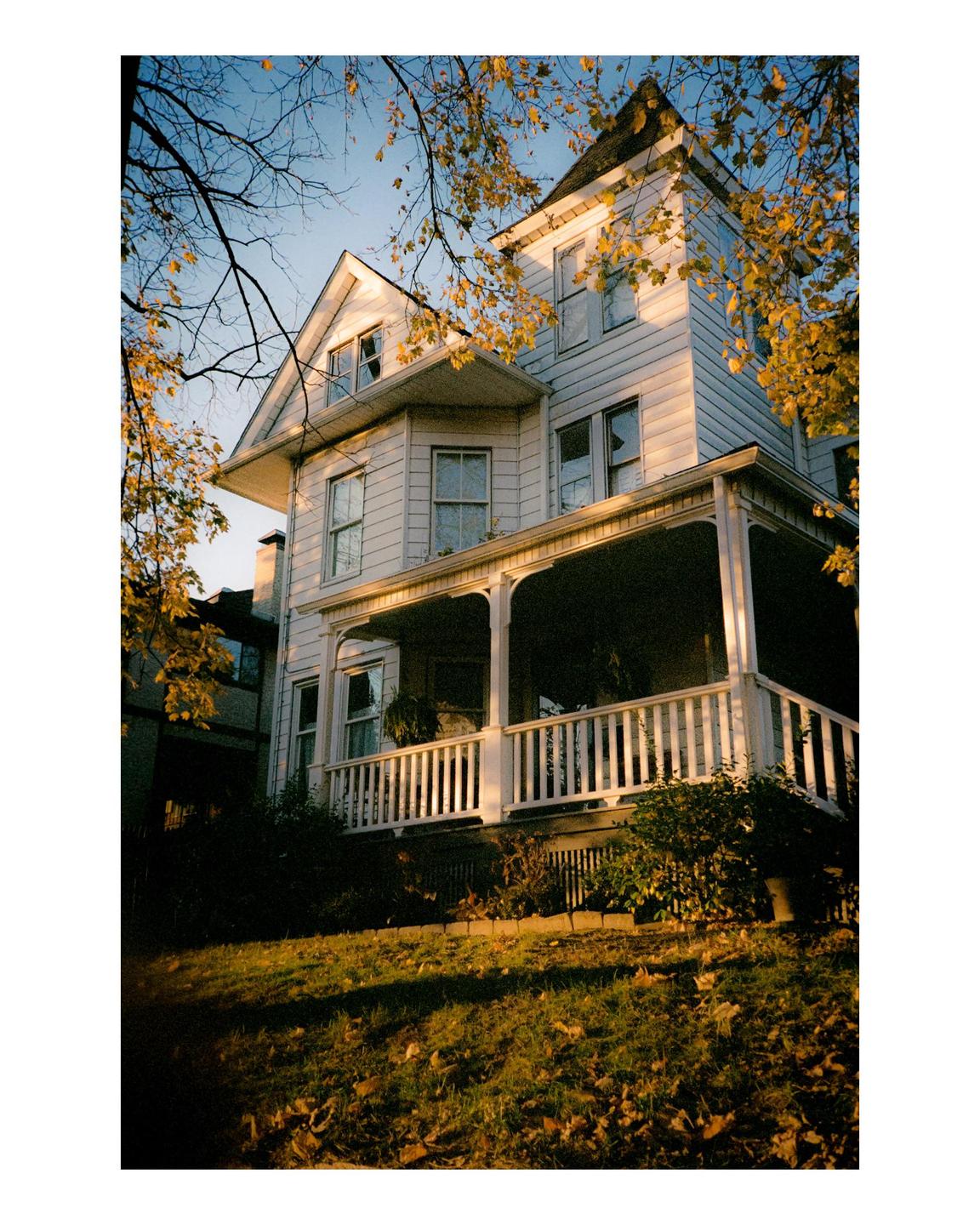 A white house with a porch is surrounded by trees with autumn foliage under a clear sky.
