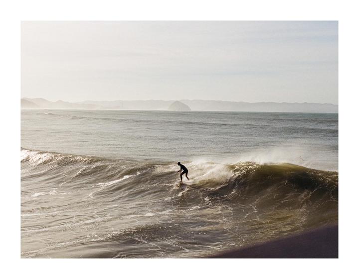 A surfer rides a wave with mountains in the background.