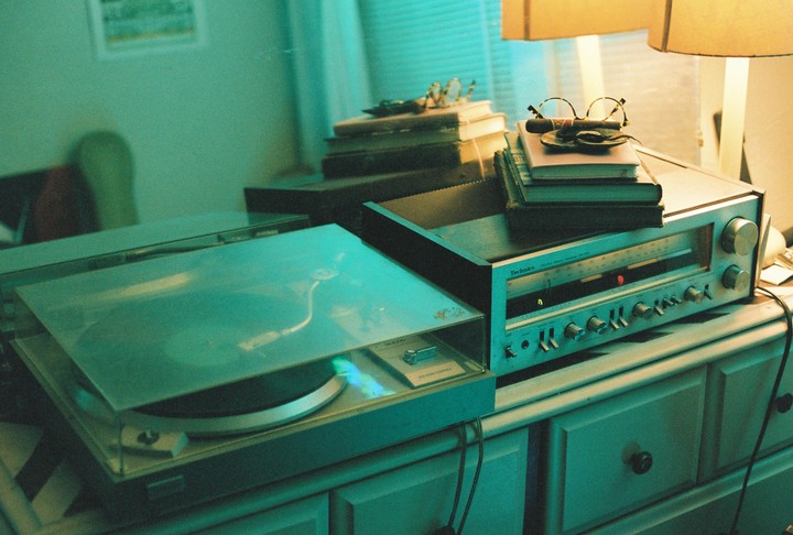 A turntable and a stereo receiver sit on a dresser next to stacks of books, with a pair of glasses and a cigar on top of one stack.