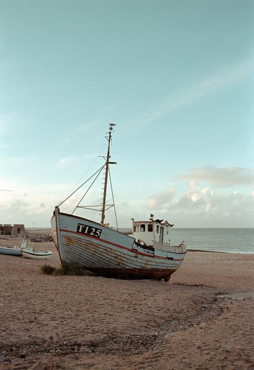 A fishing boat with the number T125 on its hull sits on a sandy beach next to the ocean under a cloudy sky.