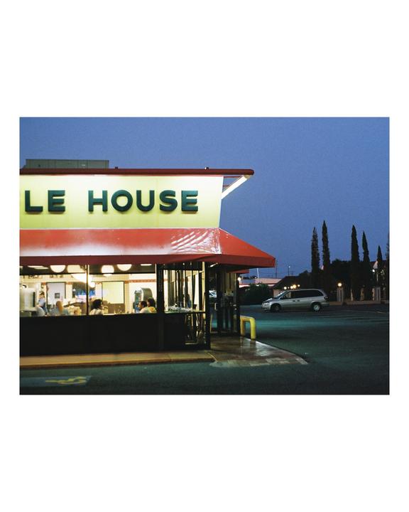 A diner with a red awning and a sign that reads "LE HOUSE" is illuminated at dusk, with a car parked outside.