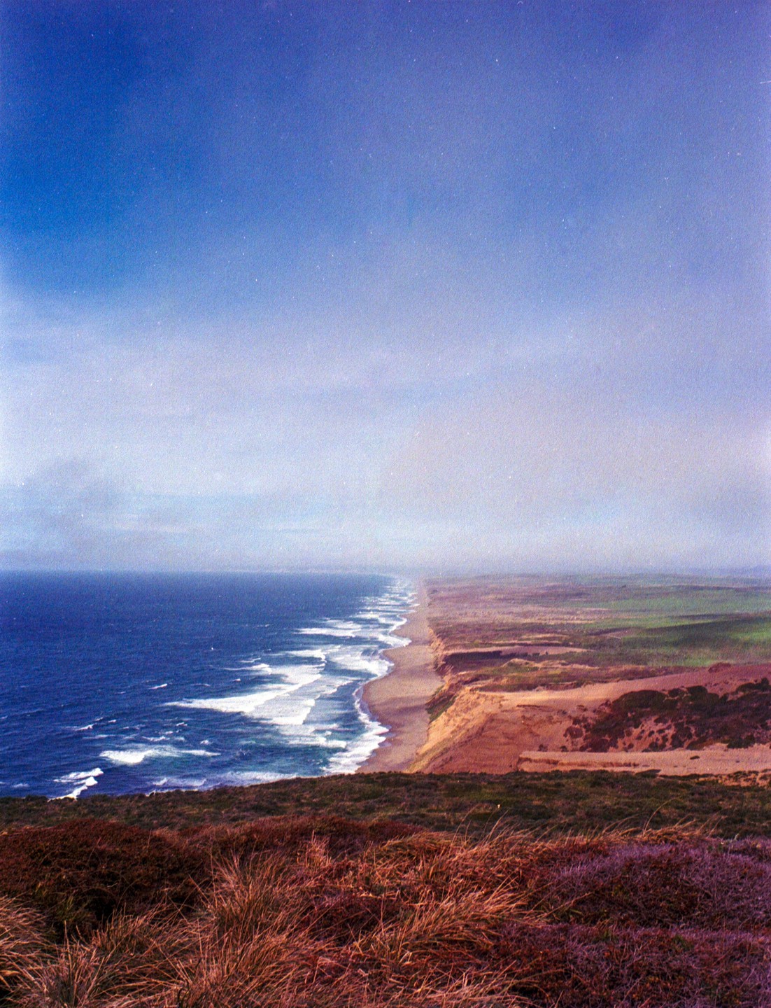 A long stretch of beach and ocean with waves crashing on the shore is seen from a high vantage point.