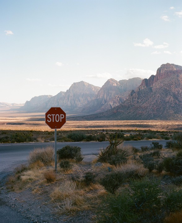 A stop sign stands at the side of a road with a vast desert landscape and mountains in the background.