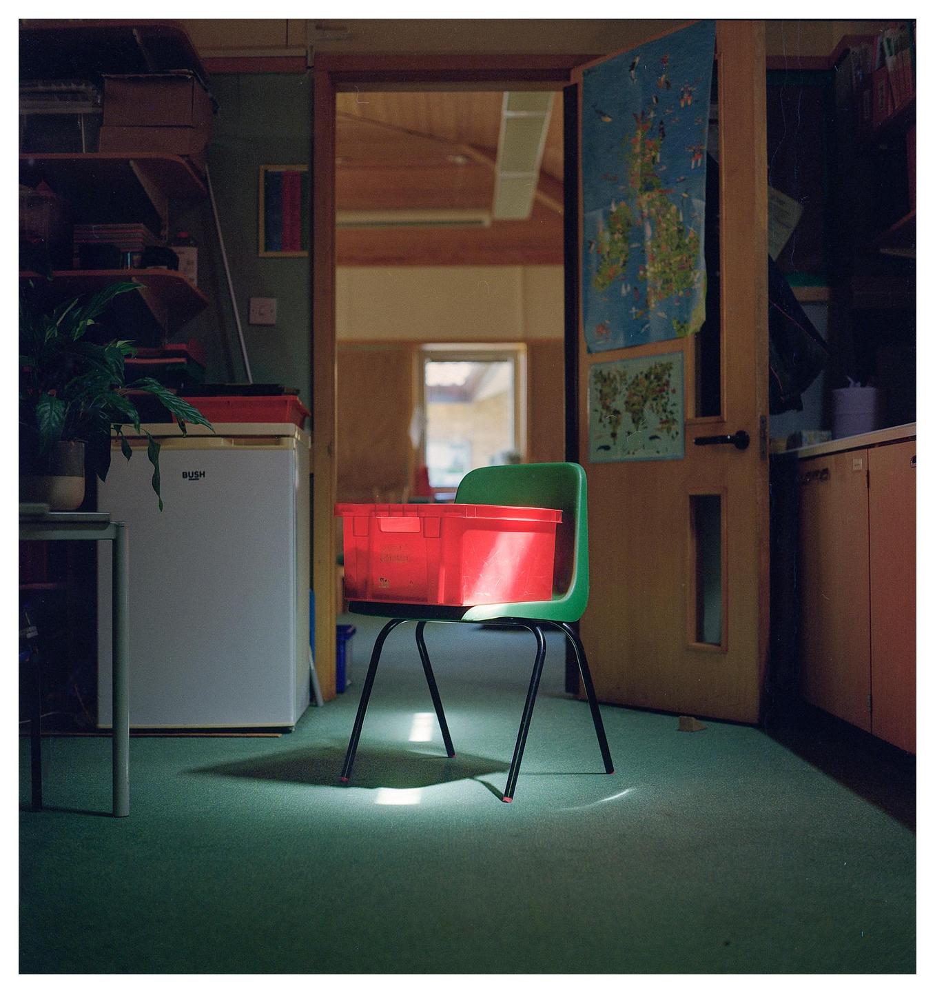 A red plastic crate sits on a green chair in a room with a map hanging on a door.