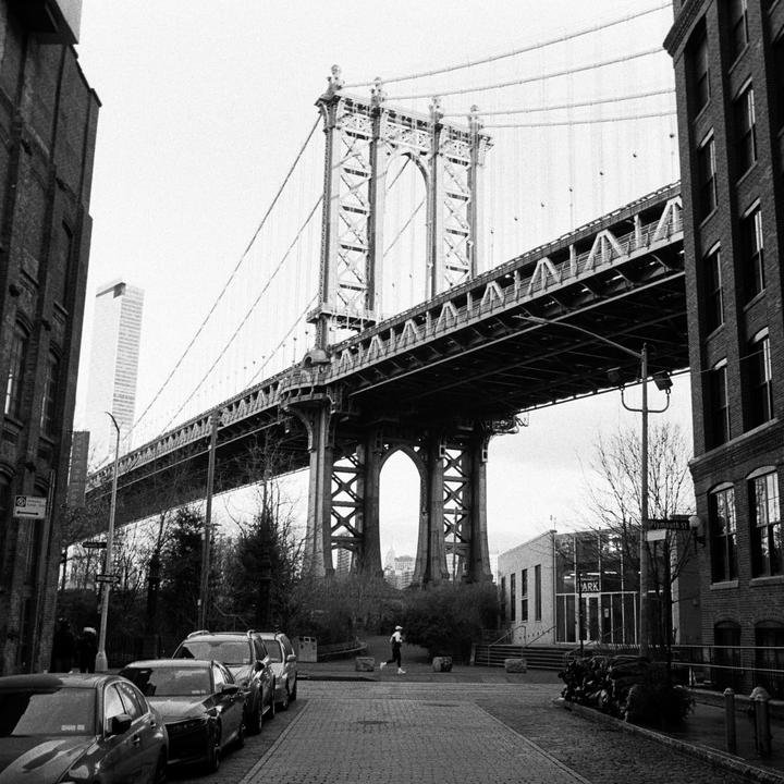 A person runs on a cobblestone street under a large bridge with cars parked on the left and buildings on either side.