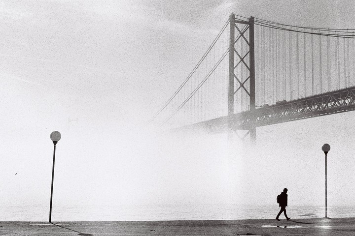 A person walks along a waterfront with a large bridge shrouded in fog in the background.