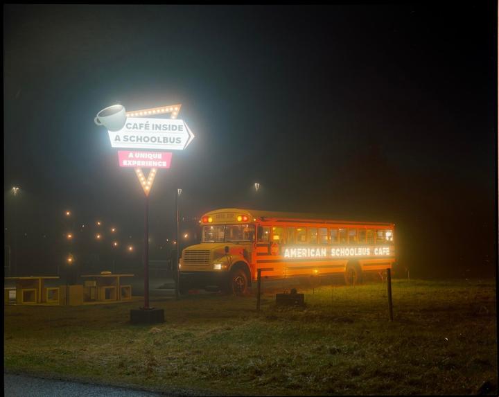 A bright yellow school bus cafe is parked in a grassy field at night, next to an illuminated sign.