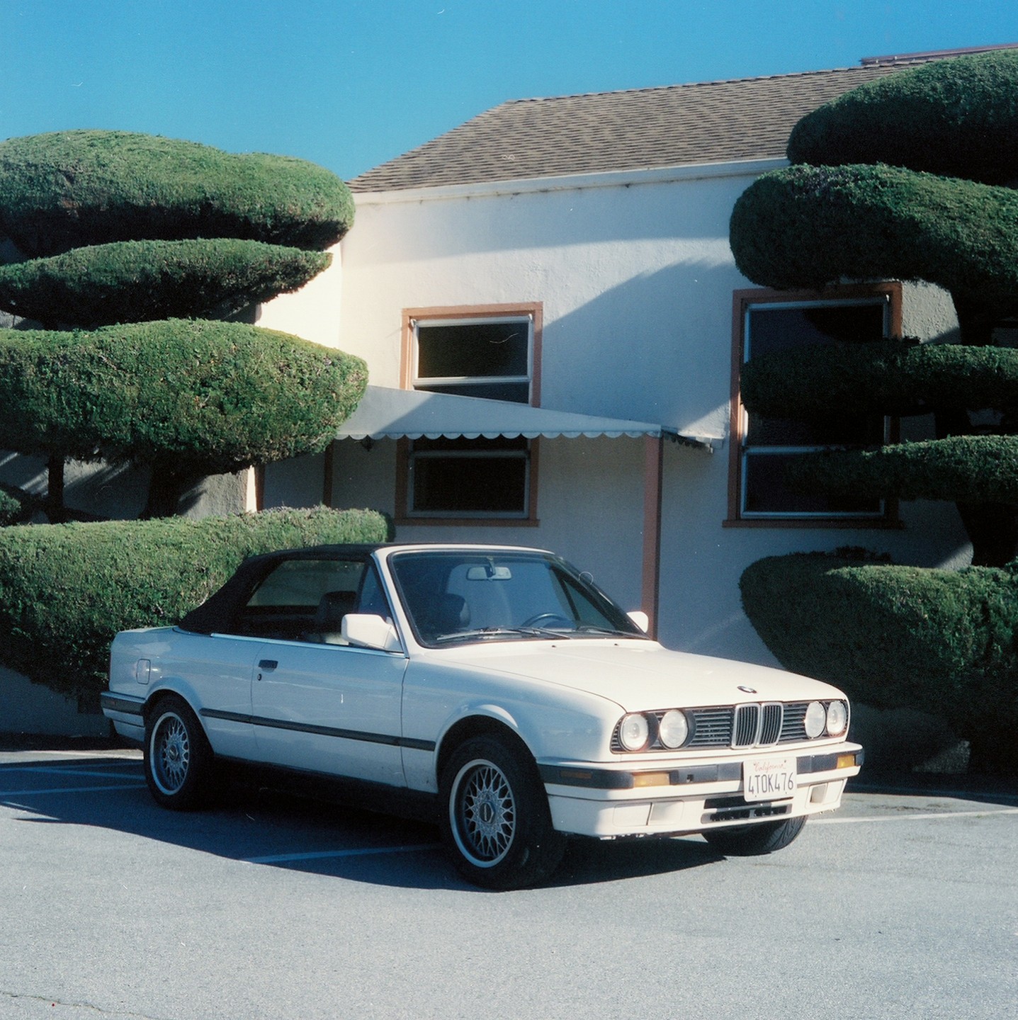 A white convertible car is parked in front of a building with sculpted bushes.