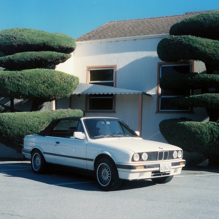 A white convertible car is parked in front of a building with sculpted bushes.