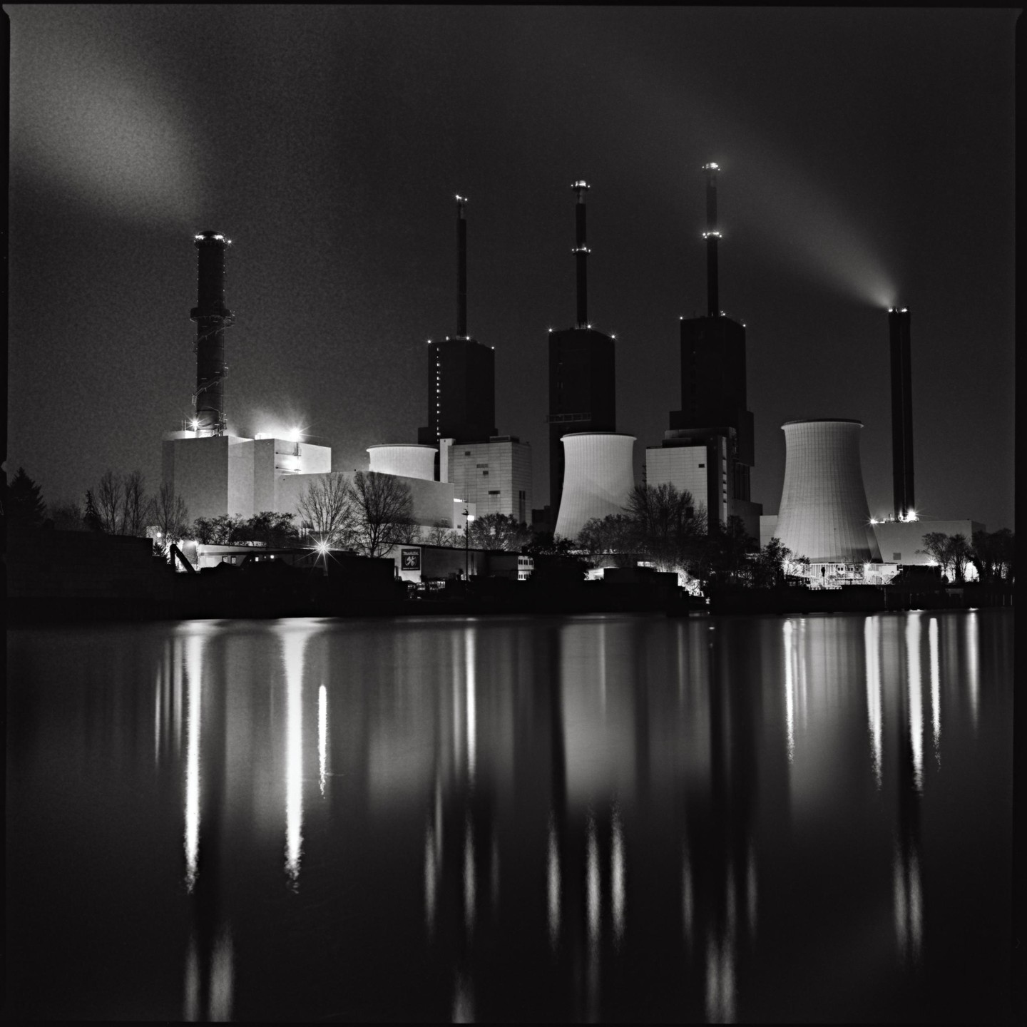 A power plant with multiple cooling towers and smokestacks is illuminated at night, reflecting in the water below.