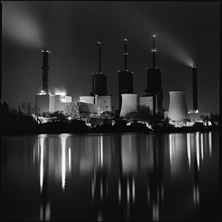 A power plant with multiple cooling towers and smokestacks is illuminated at night, reflecting in the water below.