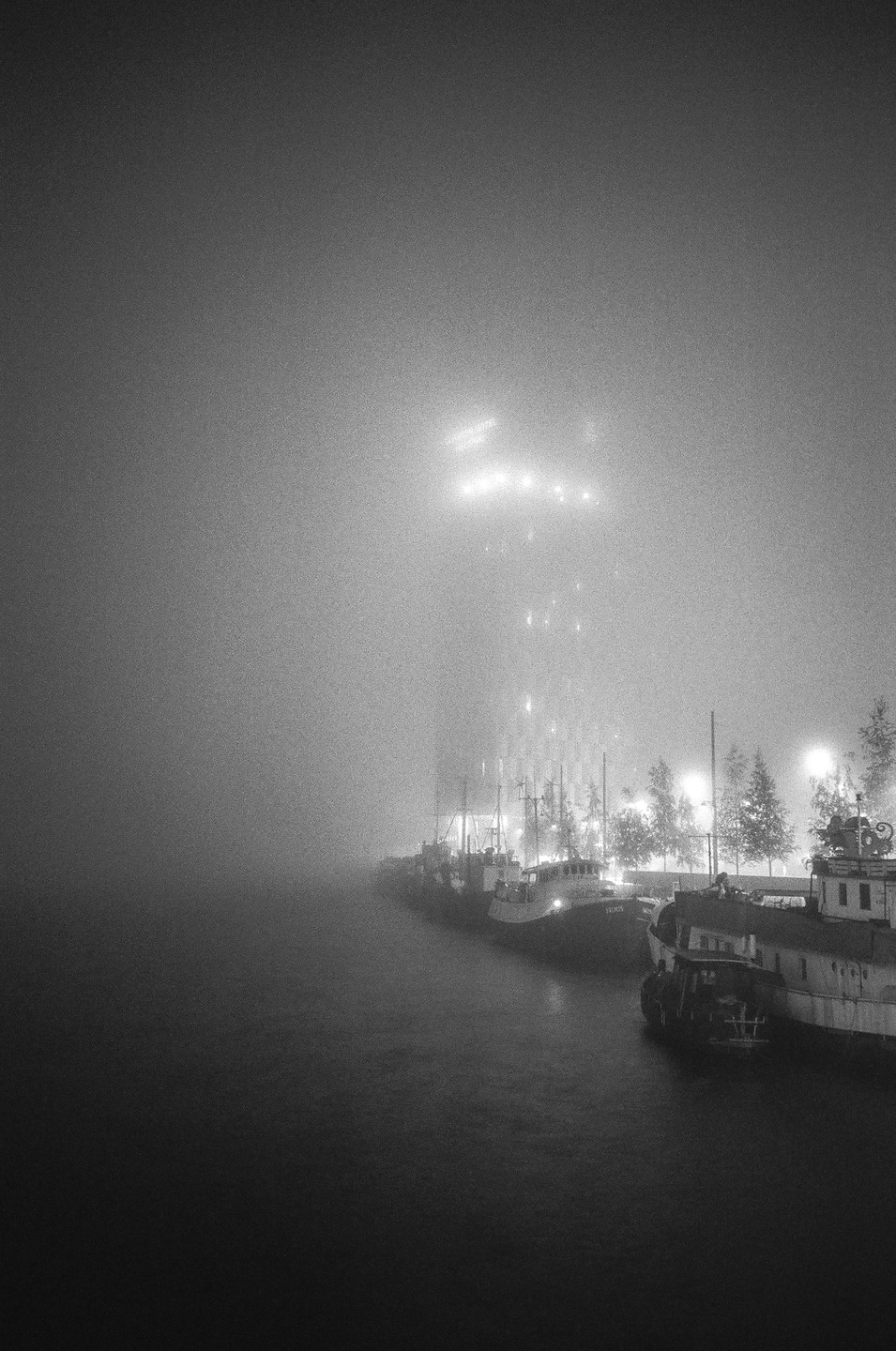 Boats are docked along a foggy waterfront at night, with a tall building visible in the distance.