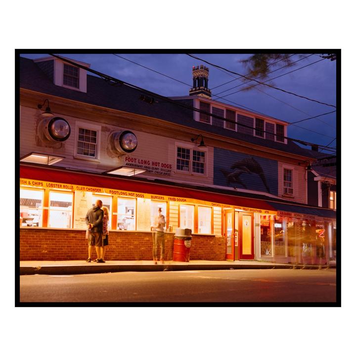 A group of people stand outside a brightly lit hot dog stand at night.