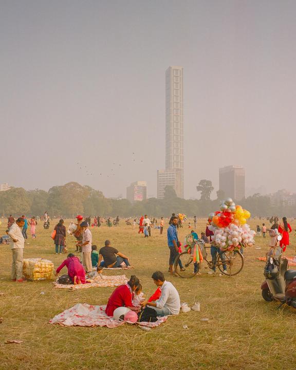 Many people are picnicking in a large grassy park with a tall building in the background.