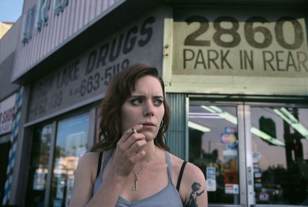 A woman with a tattoo on her arm holds a cigarette in front of a building with a "DRUGS" sign.