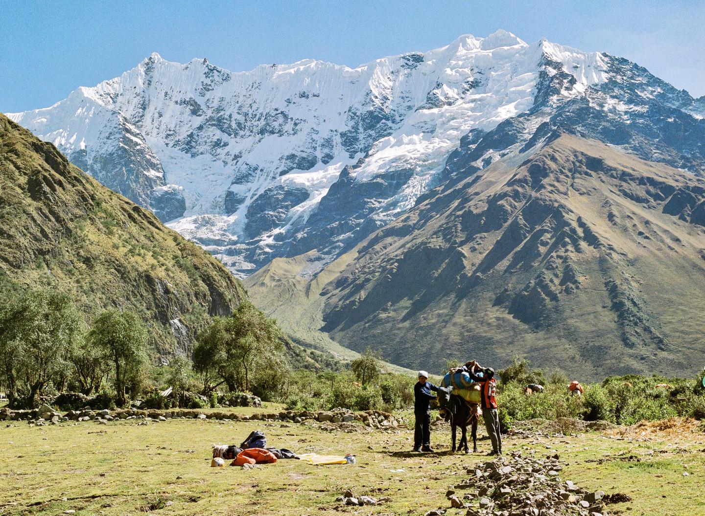 Two men load a pack animal in a mountain valley with snow-capped peaks in the background.
