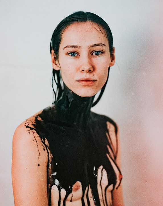 A woman with wet hair and black paint dripping down her chest looks directly at the viewer.
