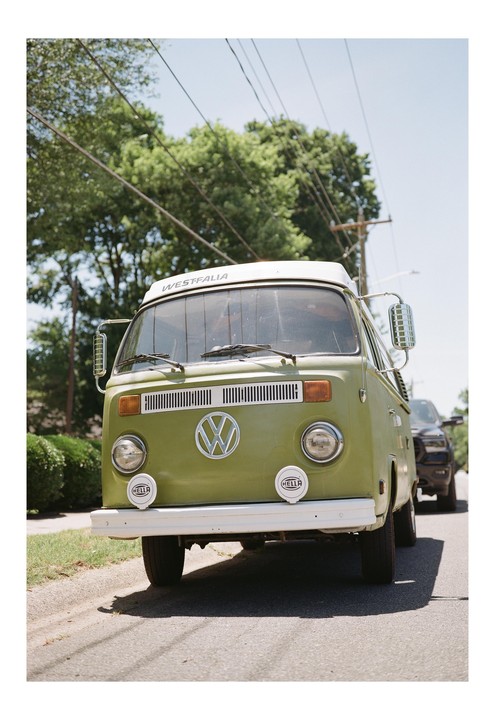 A green Volkswagen van is parked on the side of a street with trees and power lines in the background.
