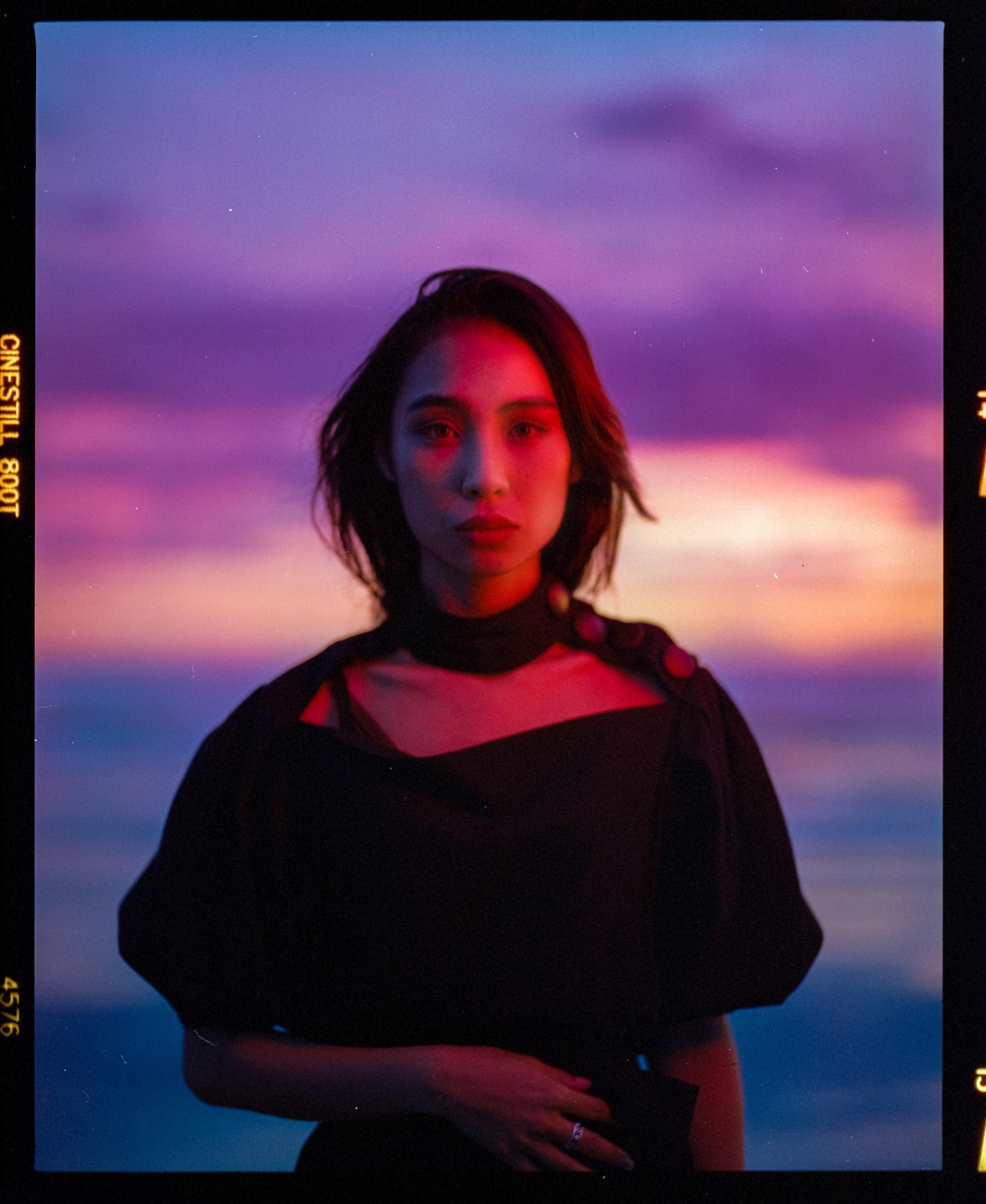 A woman with short hair and a dark top stands against a vibrant purple and orange sky.