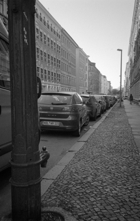 A street with parked cars and tall buildings is seen from behind a lamppost.