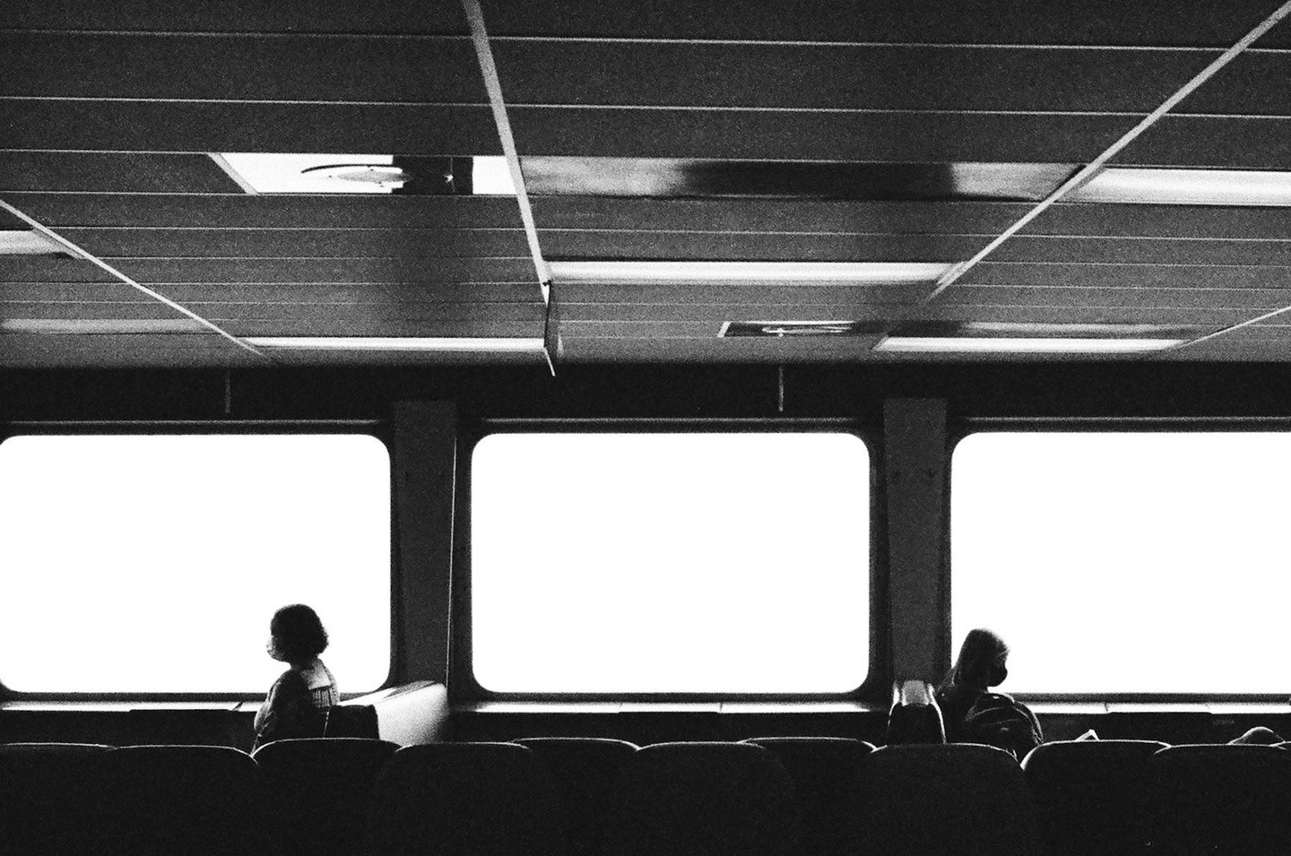 Two people sit in silhouette looking out of bright windows on a ferry.