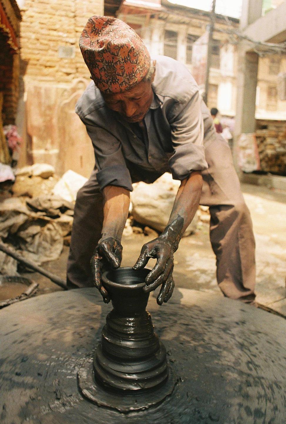 A man wearing a patterned hat works on a pottery wheel, shaping a clay pot with his hands.