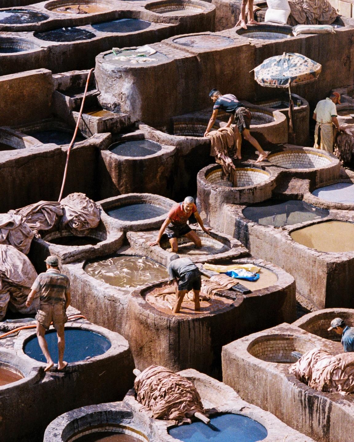 Several men work in a tannery with many stone vats filled with different colored liquids and hides.