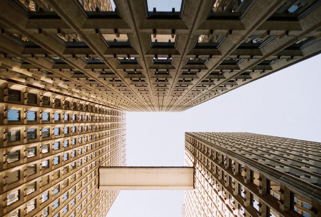 Four brutalist skyscrapers with a skybridge between two of them are viewed from directly below, looking up at the sky.