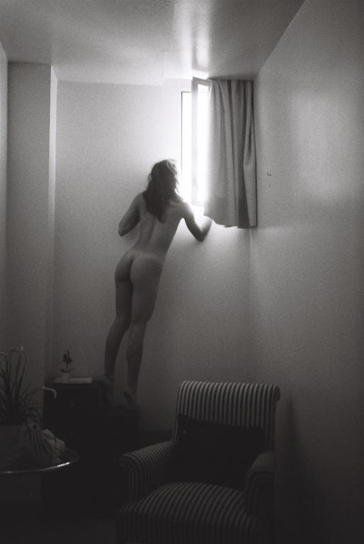 A nude woman stands on a piece of furniture to look out a window in a room with a striped armchair.
