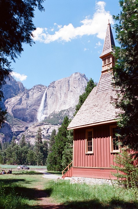 A red chapel with a tall steeple stands in a grassy field with a large waterfall and mountain in the background.